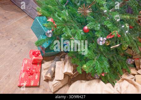 Un'accogliente scena natalizia caratterizzata da un albero sempreverde decorato con ornamenti colorati, circondato da regali avvolti e legna da ardere. Il pavimento in legno aggiunge W Foto Stock