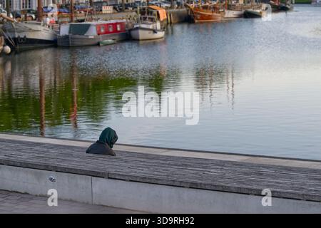 Lunghe panche in legno a più livelli vicino al porto con una persona in giacca nera cappuccio verde e cuffie sedute. La scena mostra acque calme con varie barche Foto Stock