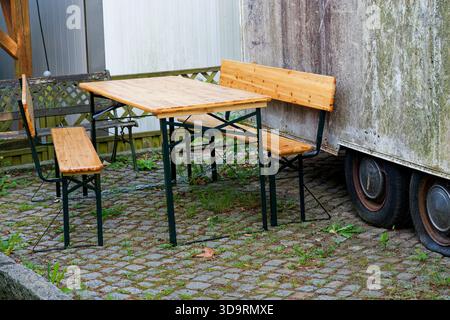 Un vecchio rimorchio con pneumatici sgonfi è macchiato di muschio e polvere. Accanto ad esso un nuovo set esterno di panche e un tavolo con gambe in metallo e spranghe in legno Foto Stock