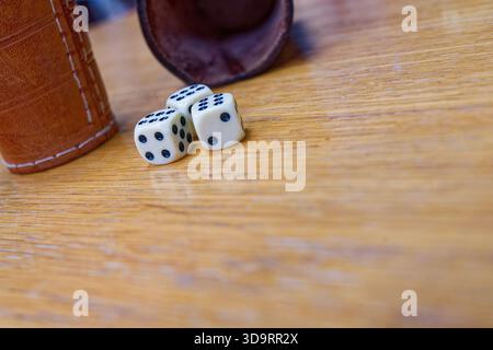 Un paio di coppe di dadi in pelle poggiano sul tavolo su entrambi i lati. Nelle vicinanze, tre dadi bianchi con picchetti neri suggeriscono una pausa durante il gioco. La superficie risplende Foto Stock