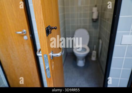 Un bagno rustico all'aperto si erge con la sua porta di legno aperta che mostra un semplice WC bianco all'interno. Un fermo metallico e un pannello ventilato sono visibili contro Foto Stock