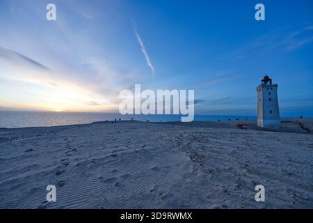 Una tranquilla spiaggia al tramonto con un faro Lyngvig Fyr. Il cielo è dipinto con sfumature blu e arancio mentre il sole tramonta all'orizzonte. S Foto Stock