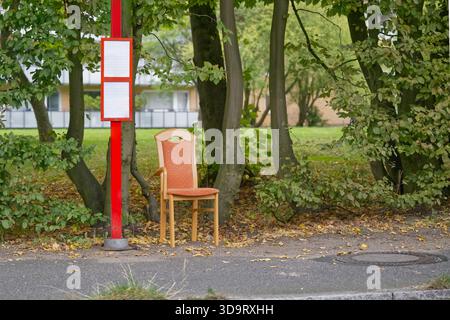 Una solitaria sedia di legno con un cuscino arancione è collocata accanto ad un cartello rosso della fermata dell'autobus, circondata da lussureggianti alberi verdi e foglie cadute a terra. Foto Stock