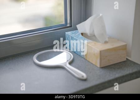 Vista ravvicinata della soglia della finestra con specchio a specchio rotondo e scatola decorativa in tessuto. Lo specchio ha una maniglia bianca, e la scatola di tessuto i Foto Stock