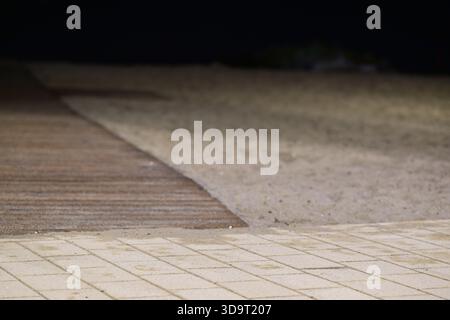 Una scena poco illuminata caratterizzata da una spiaggia di sabbia con una passerella di legno. Il terreno è un mix di piastrelle e sabbia, creando un'atmosfera tranquilla di notte al W. Foto Stock