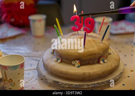Una torta di compleanno con il numero 39 in cima, circondata da candele colorate. La torta è decorata con piccole figure arcobaleno e unicorno, set o Foto Stock