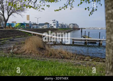 Un tranquillo panorama sul lungomare caratterizzato da un molo in legno che si estende in acque calme, circondato da banchi erbosi e da edifici moderni sullo sfondo in OLP Foto Stock