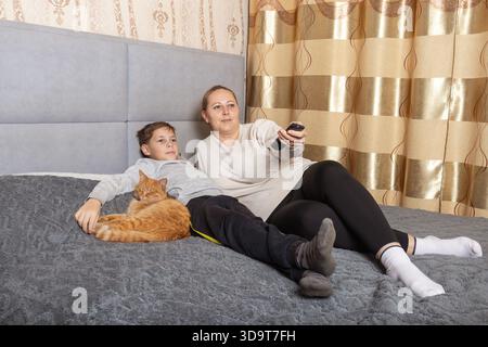 Madre e figlio si rilassano a letto con il gatto dello zenzero, guardando la televisione. per temi di legame familiare, tempo libero e comfort domestico. Foto Stock