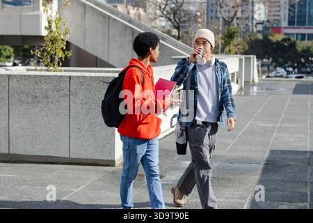 Diversi studenti maschi che camminano per la piazza sorseggiando un caffè, trasportando notebook smartphone zaini Foto Stock