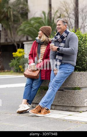 La coppia senior seduto sulla panchina della piantatrice in cemento in piazza regge tazze da caffè e indossa uno smartwatch Foto Stock