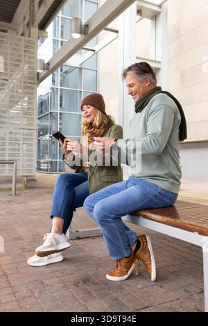 La coppia senior sedeva su una panchina di legno in piazza a controllare gli smartphone tramite pannelli di vetro e travi di metallo Foto Stock