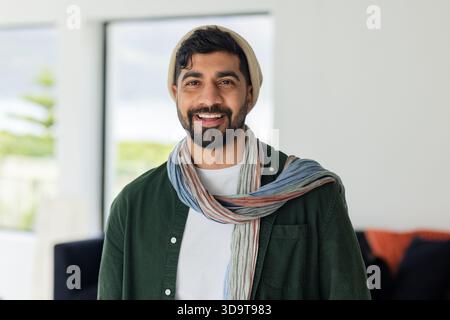 Uomo indiano in piedi in un luminoso soggiorno con berretto beige e sciarpa a righe vicino alla finestra Foto Stock