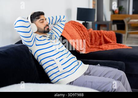 Uomo indiano sdraiato sul divano drappeggiato a casa ascoltando tramite cuffie bianche, spazio copia Foto Stock