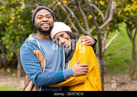 Coppia variegata che si abbraccia e sorride mentre si trova in un frutteto con beanies e sciarpa Foto Stock