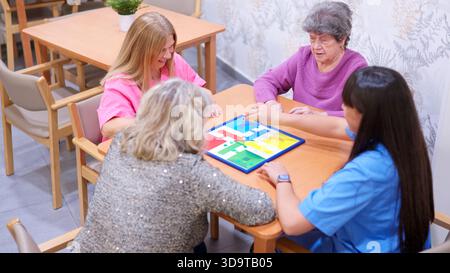 Le donne giocano a un gioco da tavolo in casa di cura con l'assistente Foto Stock