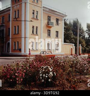 Fotografia d'archivio d'epoca degli anni '1980 che cattura un incrocio stradale affollato nel centro di Sloviansk, URSS (RSS Ucraina). Una berlina sovietica verde scuro passa davanti a un edificio residenziale a quattro piani tipico dell'architettura dell'epoca stalinista. L'angolo tra Lenin Street (ora Universitetska) e Iskra Street (ora Shovkovychna) è pieno di pedoni che attraversano la strada. Lussureggianti alberi verdi fiancheggiano i marciapiedi, ombreggiando le vetrine del negozio al piano terra. Questa immagine autentica documenta il paesaggio urbano e la tranquilla vita quotidiana nella regione del Donbas. Foto Stock