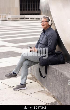 Uomo maturo seduto sulla panchina in piazza urbana a controllare lo smartphone vicino a sculture di metallo, spazio di copia Foto Stock