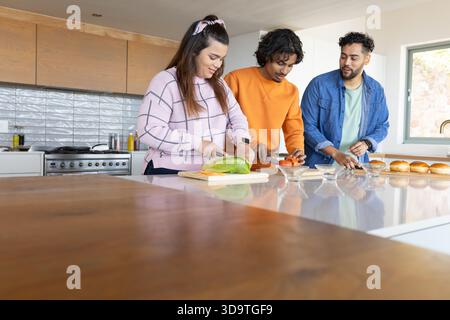 Diversi amici preparano panini all'isola della cucina di casa con lattuga, pomodori e panini Foto Stock