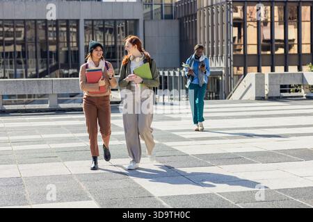 Diverse amiche che camminano sul quad del campus portando cartelle e zaini, controllando lo smartphone Foto Stock