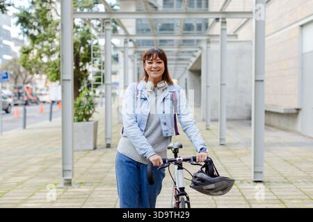 Donna sorridente che tiene in mano la bicicletta indossando cuffie e zaino sotto il pergolato di metallo sul passaggio pedonale Foto Stock