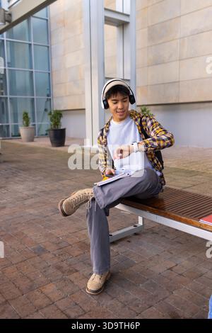 Uomo asiatico seduto sulla panchina in piazza con le cuffie che bilanciano notebook e smartphone Foto Stock