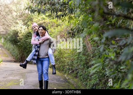 Coppia variegata che cammina lungo un sentiero boscoso che porta donna sul retro con giacca trapuntata rosa e berretto Foto Stock