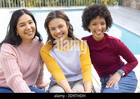 Diverse amiche che sorridono e chiacchierano mentre si siedono sulla terrazza della piscina sul retro vicino alla piscina Foto Stock