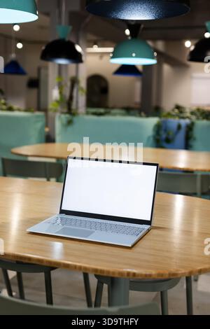 Il computer portatile sottile color argento è seduto su un tavolo ovale in legno in uno spazio di coworking open space con luci color tè Foto Stock