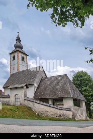 Chiesa di San Giovanni Battista a Bohinj, Slovenia, caratterizzata da un'architettura alpina storica con muri in pietra e tetti a tegole in un ambiente rurale panoramico. Foto Stock