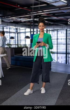 Colleghi diversi che passano accanto alla sala d'ufficio mentre uno indossa un blazer verde e utilizza lo smartphone Foto Stock