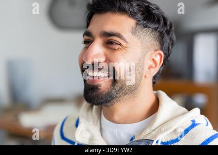 Uomo indiano che indossa un maglione bianco con cappuccio e strisce blu sorridenti in un ufficio aperto dietro la scrivania Foto Stock