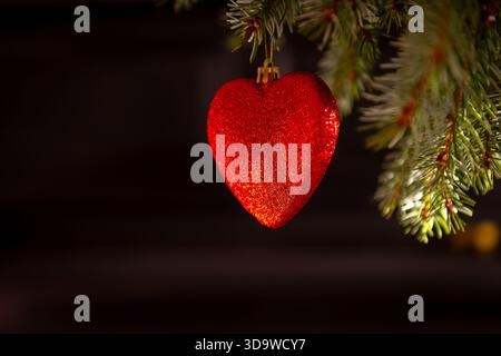 Un primo piano di una pallina di Natale rossa a forma di cuore appesa su un albero di Natale su uno sfondo scuro. Uno sfondo di elementi natalizi. Foto Stock