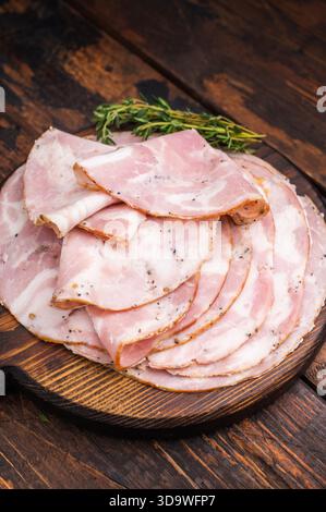 Fette di autentica pancetta arrotolata italiana servite su un tagliere rustico scuro con timo fresco e grani di pepe Foto Stock