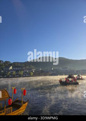 Tranquillo paesaggio mattutino del villaggio sul lago nebbioso. la barca tradizionale con lanterne rosse galleggia sull'acqua all'alba, creando montagne e cielo limpido Foto Stock