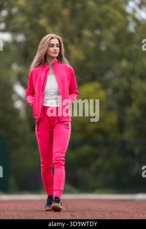 una giovane donna cammina su una pista da running indossando un abito luminoso. Il sole splende su di lei mentre passeggia attraverso un parco pieno di alberi in una giornata limpida. Foto Stock