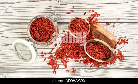 Goji secco, aka. Semi di mirtillo all'interno di una bottiglia di vetro, alcuni in ciotole di legno e versato sulla scrivania delle tavole bianche, vista dall'alto Foto Stock