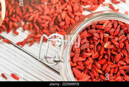 Goji secco aka. Semi di mirtillo all'interno di una bottiglia di vetro, alcuni sparsi su una scrivania di tavole bianche Foto Stock
