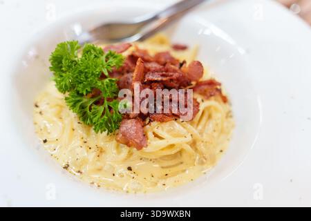 Primo piano di cremosi spaghetti alla carbonara serviti con pancetta croccante e prezzemolo fresco in una ciotola bianca presso il ristorante italiano Foto Stock
