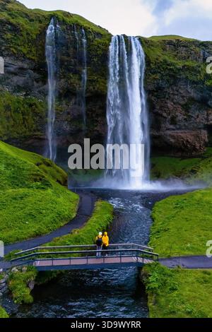 I visitatori si trovano su un ponte presso la cascata Seljalandsfoss, circondati da vegetazione lussureggiante e scogliere spettacolari in Islanda. L'ambiente tranquillo cattura l'essenza dello splendore della natura. || modello rilasciato Foto Stock