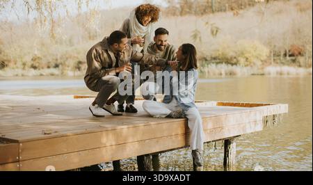 Un gruppo di quattro amici condivide momenti di gioia su un molo in riva al lago, sorseggiando bevande e ridendo, circondati dalla natura alla luce calda del sole. Foto Stock