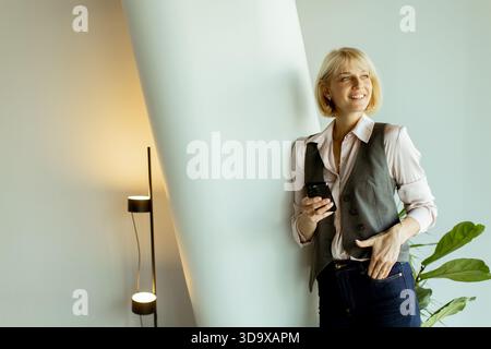 Una donna con i capelli biondi corti si appoggia contro una parete in un ufficio elegante. Sorride e tiene in mano uno smartphone, vestito con un gilet grigio e un shi leggero Foto Stock