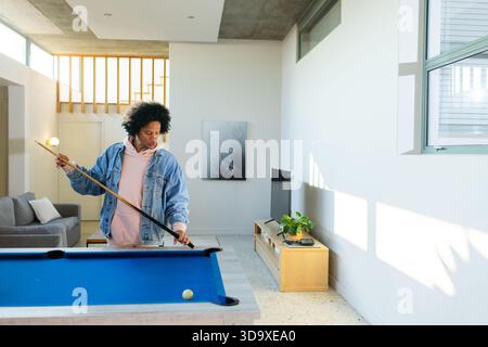 Uomo afro-americano che punta il bastone al tavolo da biliardo blu in soggiorno, spazio copia Foto Stock