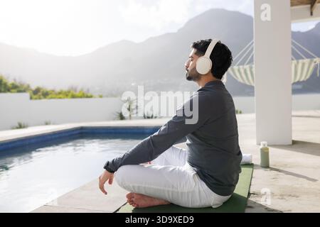 Uomo seduto su un tappetino verde per yoga vicino alla piscina con una bottiglia sportiva e indossa cuffie vicino alla valle della montagna Foto Stock
