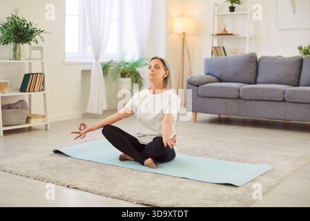 Donna matura seduta con le gambe incrociate sul tappetino, meditando a occhi chiusi, facendo esercizi di respirazione. Foto Stock