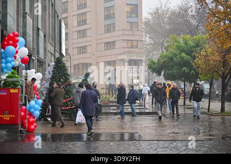 Erevan Armenia 12.7,2025. Un gruppo di persone sta camminando lungo una strada bagnata di fronte a un edificio. La strada è coperta di pozzanghere e la gente lo è Foto Stock