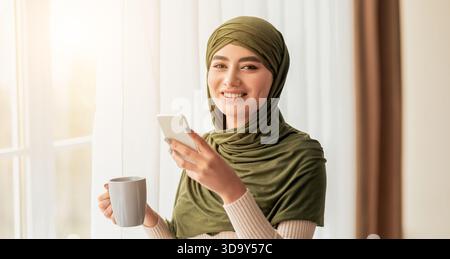 Giovane donna che si gusta un caffè mentre usa uno smartphone vicino a una finestra Foto Stock