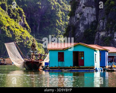 Barca in legno e case di pescatori colorate su zattere galleggianti adagiate contro spettacolari formazioni carsiche di pietra calcarea e acque color smeraldo della baia di ha Long, Vietnam Foto Stock