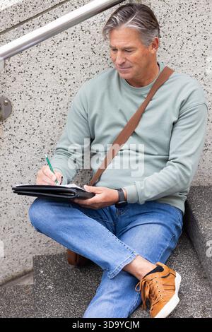 Uomo anziano che scrive su un notebook usando una penna verde su gradini esterni con corrimano e borsa Messenger Foto Stock