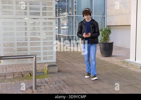 Uomo che cammina su asfaltatrici di mattoni accanto al pannello a strisce utilizzando smartphone e cuffie, spazio di copia Foto Stock