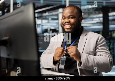 Un amministratore di sistema sorridente nel data center che controlla lo stato dell'infrastruttura, mostra indicatori di prestazioni stabili. Felice specialista di sala server euforico dopo aver completato con successo le operazioni Foto Stock